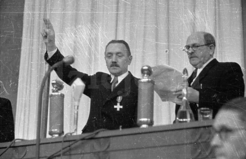 Czarno‑białe zdjęcie przedstawia dwóch mężczyzn. Obaj są ubrani w ciemne garnitury i stoją za długim stołem. Ten stojący po prawej czyta tekst z kartki i ma na sobie okulary. Ten po prawej wyciąga w górę prawą dłoń z wystawionymi dwoma palcami i patrzy na blat stołu. NA stole stoją mikrofony i szklanki z wodą. 