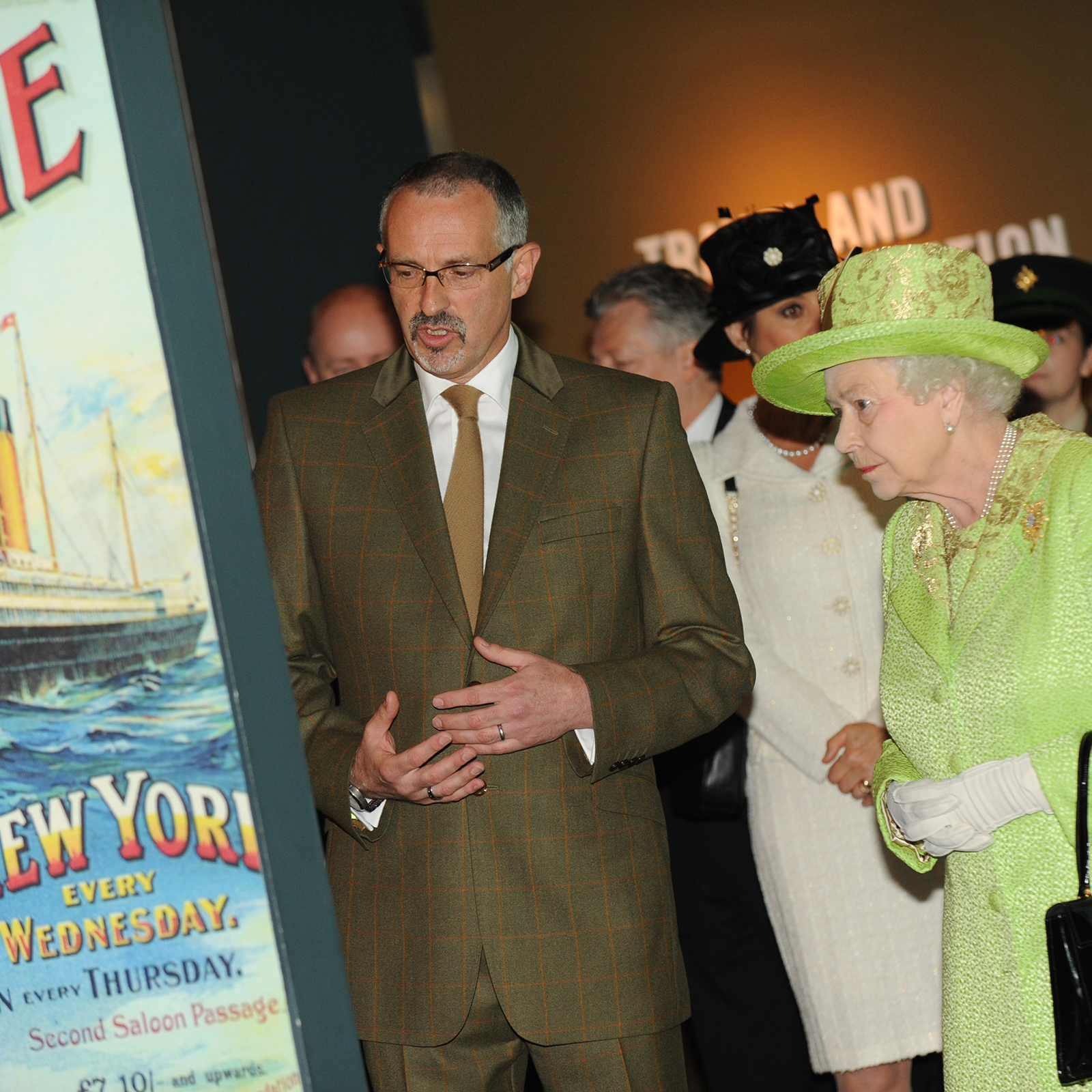 Zdjęcie przedstawia królową Elżbietę II uczestniczącą w jakimś wydarzeniu i przyglądającą się posterowi. Widoczna jest starsza kobieta, która ubrana jest w jaskrawozielony kostium, jaskrawozielony kapelusz i białe rękawiczki. Kobieta stoi zwrócona w lewo i spogląda na coś, co jest omawiane przez mężczyznę stojącego po jej prawej stronie. Mężczyzna w średnim wieku jest ubrany w garnitur w kolorze zgniłozielonym. Obie ręce ma uniesione na wysokość pasa i gestykuluje nimi. W tle widoczne są inne osoby.