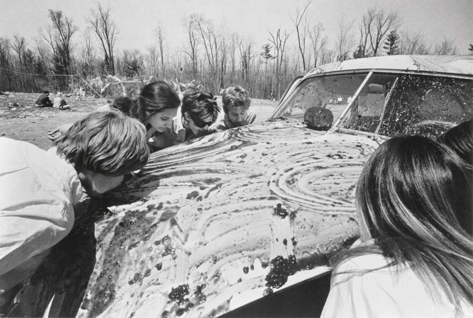 Stare czarno białe zdjęcie przedstawia auto, wokół którego zebrało się kilka kobiet. Maska pojazdu pokryta jest dżemem, który zlizywany jest przez zgromadzone osoby.
