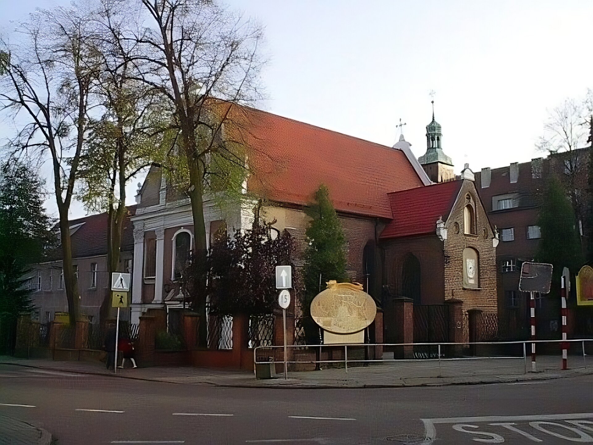 Zdjęcie współczesne przedstawia widok na zewnętrzną ścianę nawy głównej i skrzydła kościoła św. Anny w Wejherowie. Fasada kościoła w stylu barokowym, jasne fryzy, gzymsy i pilastry stanowią wykończenie frontonu kościoła o dwuspadowym dachu. W głębi zdjęcia krzyż nad prezbiterium, skrzydło konstrukcji ceglanej o dwuspadowym dachu z bocznym wejściem do kościoła. Nad wejściem widoczne dwa otwory okienne na witraże. Za prezbiterium i skrzydłem widoczny fragment wieżyczki z neogotyckim wykończeniem szczytu. W tle zabudowania domów mieszkalnych z cegły. Na pierwszym planie ulica dwupasmowa.