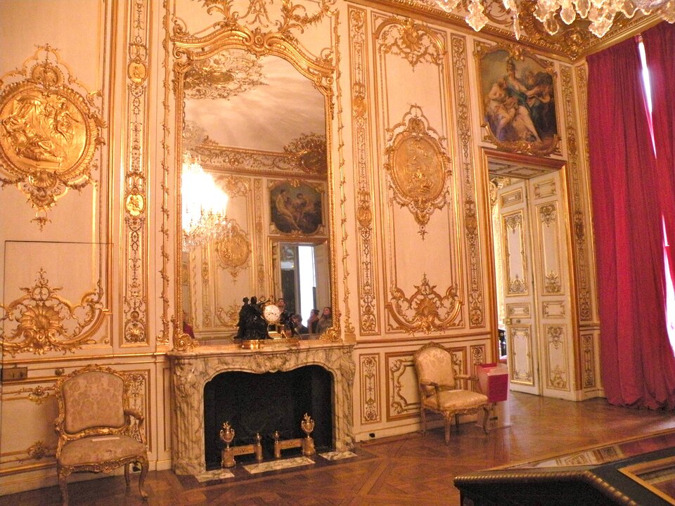 Ilustracja ukazuje wnętrze Hôtelu de Soubise w Paryżu. Ilustracja przedstawia bogato zdobione wnętrze. Ściany pokryte są licznymi ornamentami w kolorze złotym oraz obrazami w ozdobnych ramach. Widoczne jest również duże lustro w złotej ramie, które wisi nad marmurowym kominkiem. Po obu stronach kominka stoją złote, tapicerowane krzesła. Okna zasłonięte są czerwonymi zasłonami.