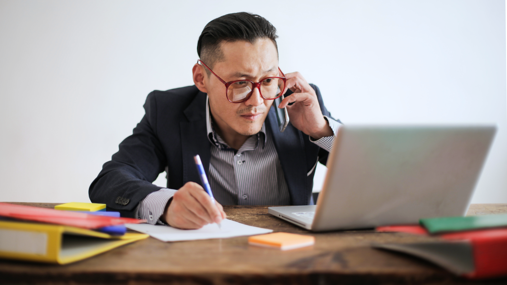 Zdjęcie przedstawia mężczyznę siedzącego przed laptopem. Mężczyzna rozmawia przez telefon i wykonuje notatki na kartce papieru.