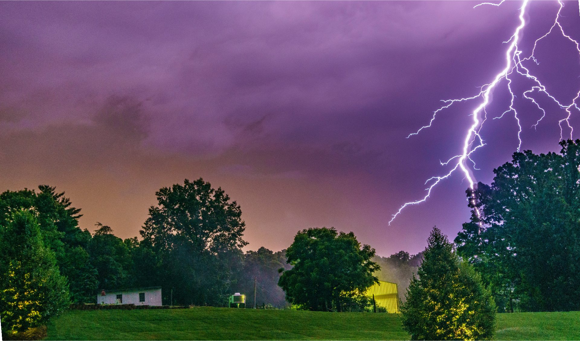Ilustracja przedstawia piorun rozświetlający łąkę, na której znajduje się domek i kilka drzew.