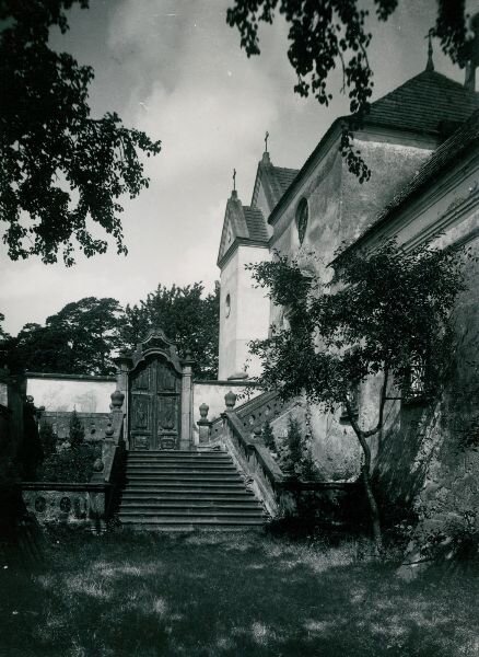 Zdjęcie przedstawia murowane schody z betonową balustradą prowadzące do bramy, za którą znajduje się kościół.