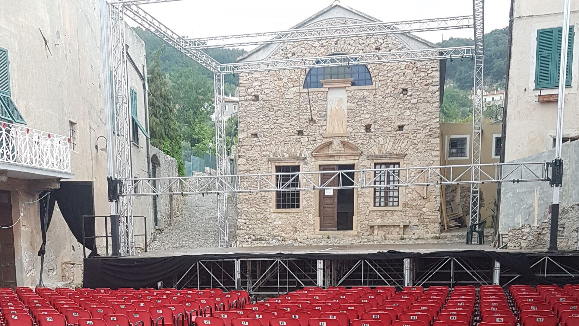 Zdjęcie przedstawia scenę teatralną w plenerze. Scena umiejscowiona jest pomiędzy starymi, kamiennymi budynkami. Przed sceną znajdują się rzędy pustych krzeseł. 