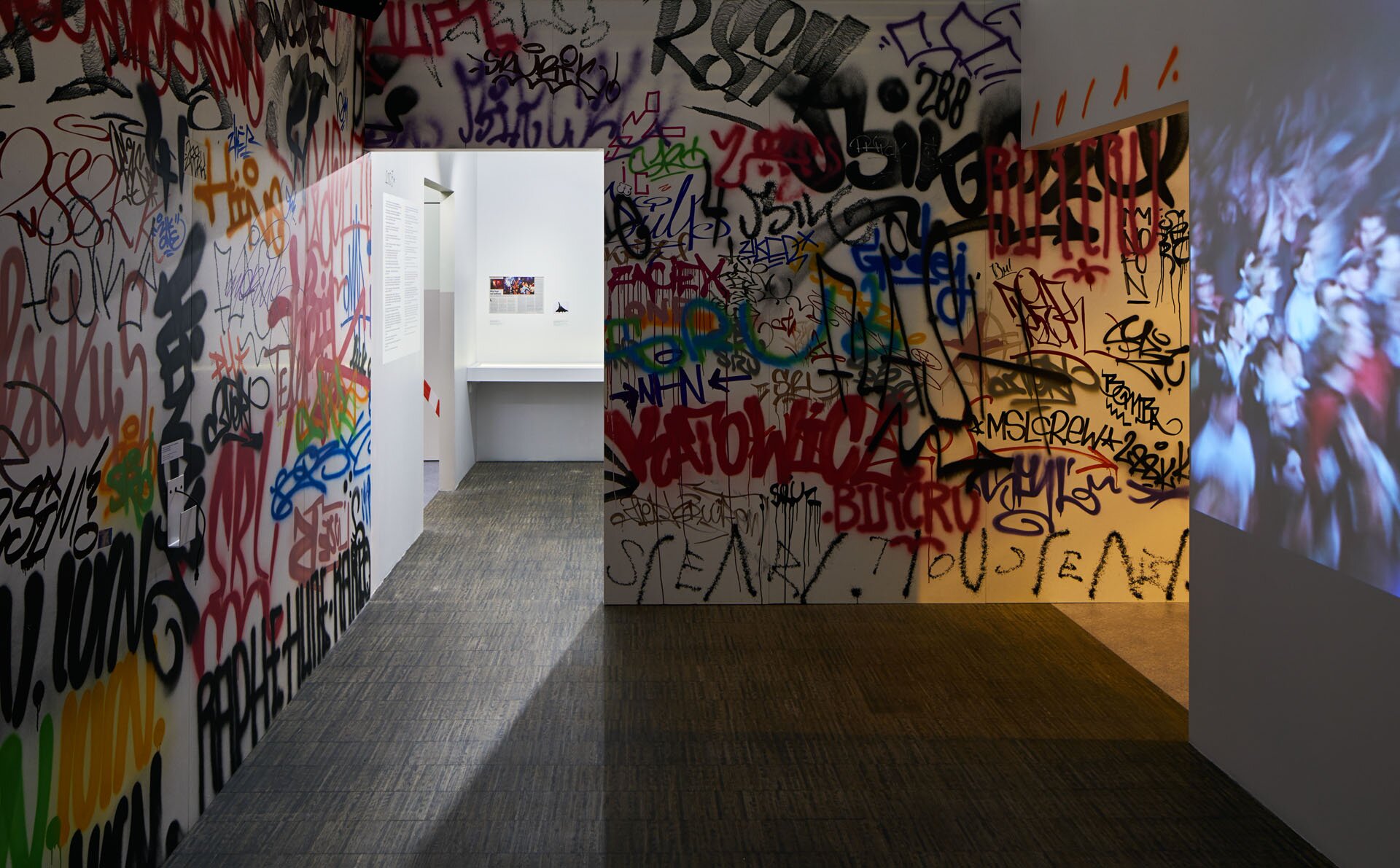 Fotografia Rafała Wyrwicha przedstawia salę z fragmentem ekspozycji pt. Zajawka. Na białych ścianach znajdują się liczne graffiti w kolorach czarnym, żółtym, czerwonym, zielonym, pomarańczowym oraz niebieskim. Napisy, z których większość jest nieczytelna, nachodzą na siebie lub dotykają się. Na ścianie z prawej strony znajduje się zdjęcie przedstawiające tłum ludzi, które zostało wyświetlone za pomocą rzutnika. Tło stanowią dalsze ściany pomieszczenia, niepokryte graffiti. 