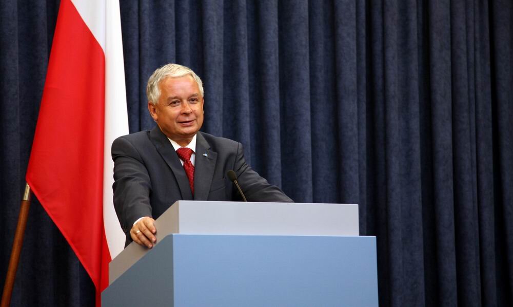Kliknij żeby powiększyć Na zdjęciu znajduje się prezydent Lech Kaczyński podczas wystąpienia na mównicy. W tle jest flaga Polski. Starszy mężczyzna z siwymi włosami ubrany w garnitur.