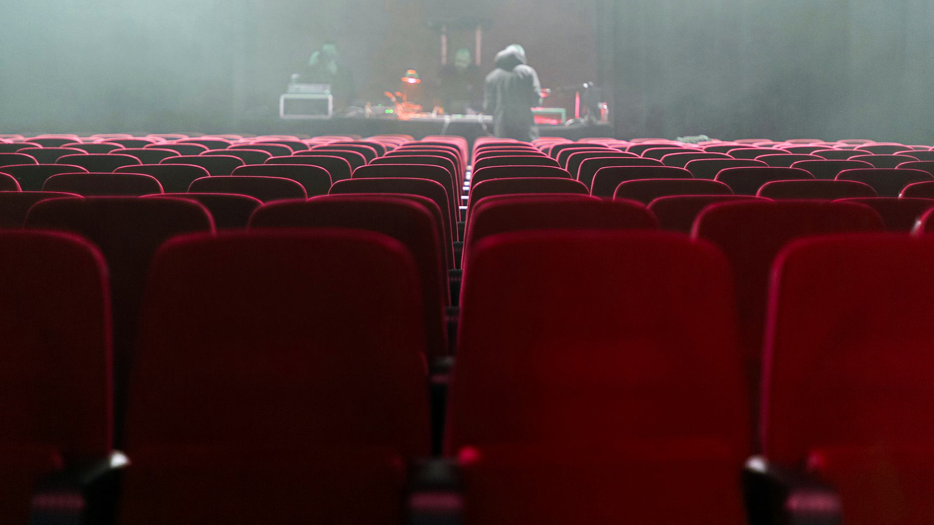 Zdjęcie przedstawia wnętrze sali kinowej z pozycji ostatniego rzędu. Na pierwszym planie widoczny jest ostatni rząd. W tle znajduje się fragment ekranu kinowego, na którym wyświetlany jest film.