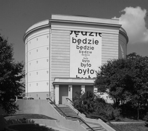 Zdjęcie przedstawia budynek. Na elewacji budynku znajduje się mural. Składa się on z napisu będzie, które jest napisane wielokrotnie jeden pod drugim. Zmniejsza się od góry do środka. Następny napis to było, które też jest napisane wielokrotnie jeden pod drugim i zwiększa się od środka do dołu.