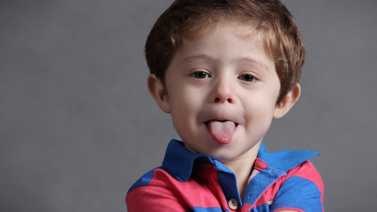 Zdjęcie przedstawia kilkuletniego chłopca, w ujęciu od ramion w górę. Chłopiec patrzy w obiektyw, ma ciemne, lekko rude, krótkie włosy, jest ubrany w koszulkę polo w czerwone i niebieskie paski. Ma zadowolony wyraz twarzy i wystawiony język.