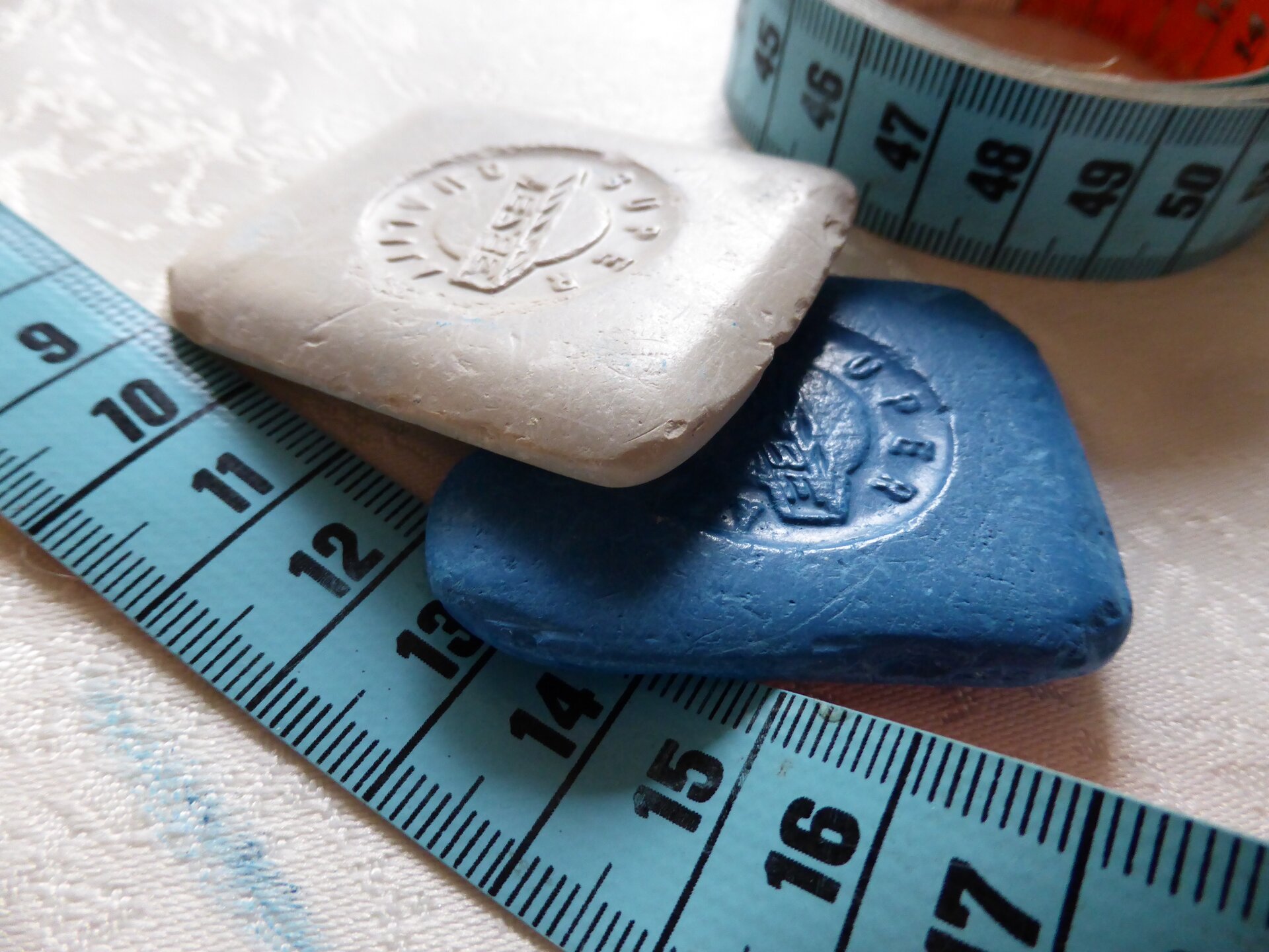 Zdjęcie przedstawia dwa centymetry krawieckie oraz dwie kwadratowe kredy krawieckie. Mają kwadratowy kształt. Jedna jest biała, a druga niebieska.