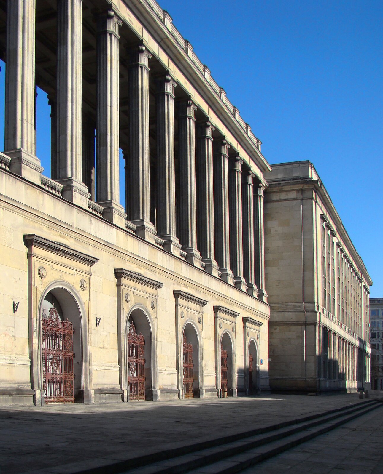 Zdjęcie przedstawia duży budynek z okazałymi, okratowanymi wejściami oraz długą kolumnadą.