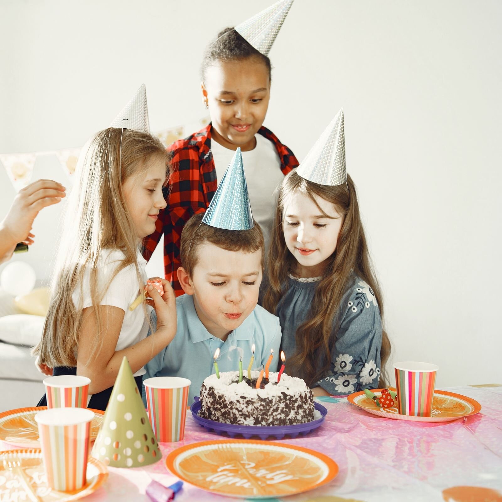 Zdjęcie przedstawia czwórkę małych dzieci. W centralnej części widoczny jest sześcioletni chłopiec siedzący przy stole, pochylający się nad stojącym na stole tortem urodzinowym. Na torcie pali się sześć kolorowych małych świeczek, a chłopiec je zdmuchuje. Wokół chłopca stoją trzy dziewczynki, które spoglądają na zdmuchiwane świeczki i tort urodzinowy. Wszyscy mają na głowach kolorowe czapeczki urodzinowe. Na stole widoczne są również kolorowe kubeczki i talerzyki oraz jedna czapeczka urodzinowa.