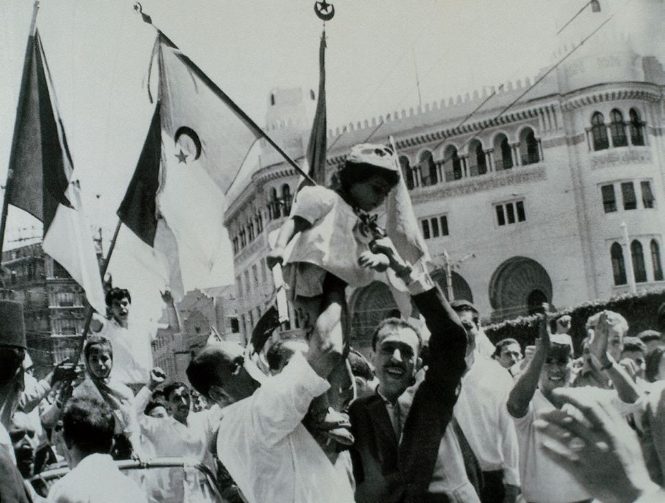 Na zdjęciu znajduje się scena podczas referendum w sprawie niepodległości Algierii, 1 lipca 1962 roku, kiedy to demonstranci z algierskimi flagami przechodzą przez dzielnicę zamieszkiwaną przez Europejczyków. Fotografia przedstawia wiwatujący tłum w białych ubraniach – ludzie śmieją się, klaszczą i krzyczą. Na pierwszym planie znajduje się dwóch mężczyzn – jeden z nich ma białą koszulę, drugi jest ubrany w ciemny garnitur. Podnoszą do góry małą dziewczynkę w białej sukience i kapelusiku. Trzymają w ręce flagi w poprzeczny pas z półksiężycem i gwiazdą. W tle widać zabudowania miasta, w tym bogato zdobioną fasadę kamienicy z półokrągłymi bramami, oknami i licznymi kolumnami. 