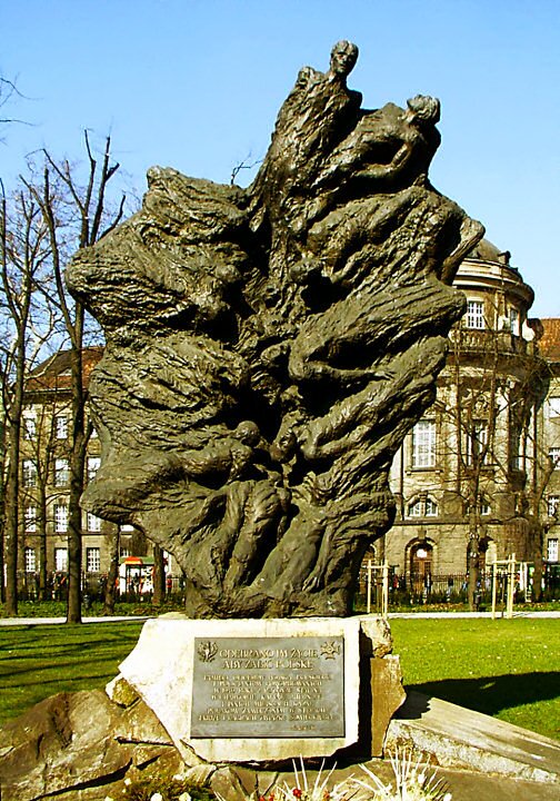 Ilustracja przedstawia pomnik w formie nieregularnej płyty, przedstawiającej ciała ludzkie wciągane do pionowej szczeliny. Widać drobne akcesoria wojskowe, miedzy innymi: szable, sprzączki od pasa itp. W tle budynek .