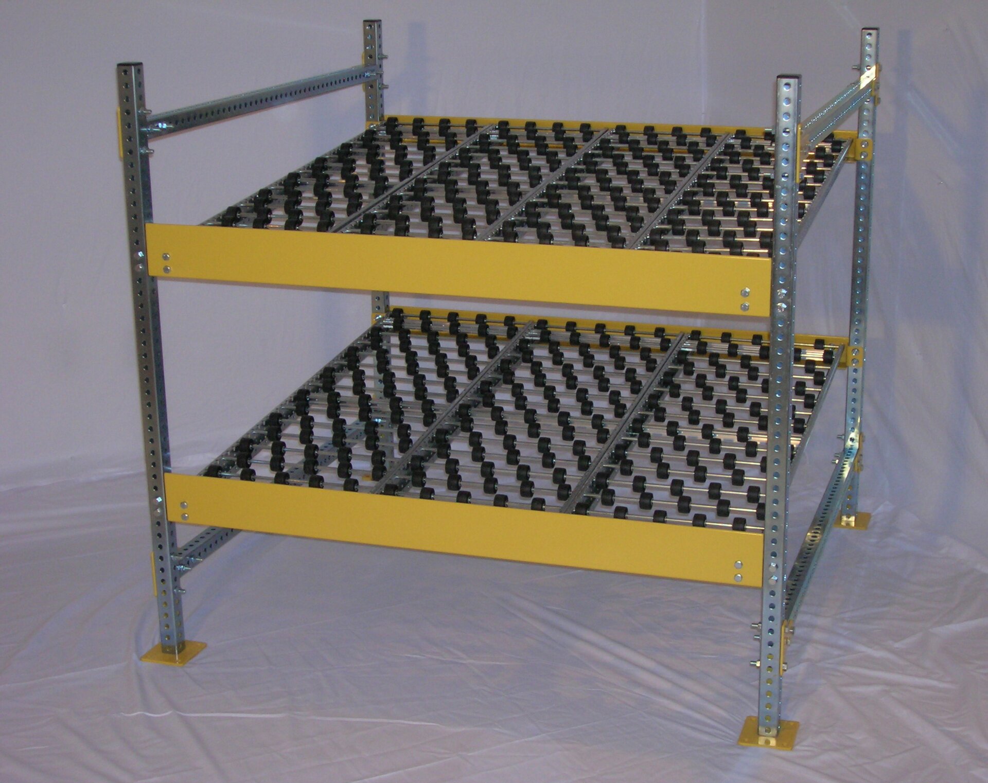 Na grafice regał przepływowy (grawitacyjny), składający się z czterech podpórek i dwóch półek.