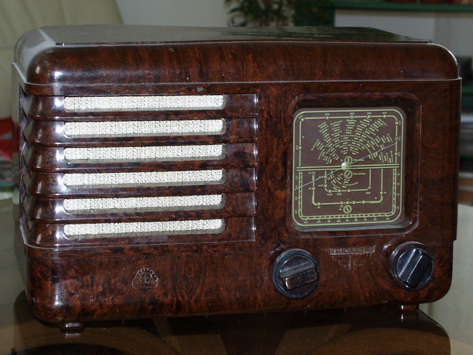 Rys. a. Ilustracja przedstawia zdjęcie, na którym widoczne jest radio. Odbiornik radiowy pochodzi z połowy dwudziestego wieku. Odbiornik radiowy widoczny jest w postaci prostopadłościennego brązowego pudełka. Obudowa wykonana jest z drewna. Na przodzie widoczny jest głośnik. Głośnik widoczny jest w postaci poziomych prostokątnych elementów w kolorze białym. Po prawej stronie widoczne są dwa czarne pokrętła. Pokrętła służą do regulacji częstotliwości odbieranej przez radio. Po prawej stronie widoczna jest tablica, na której wskaźnik pokazuje wybraną częstotliwość.