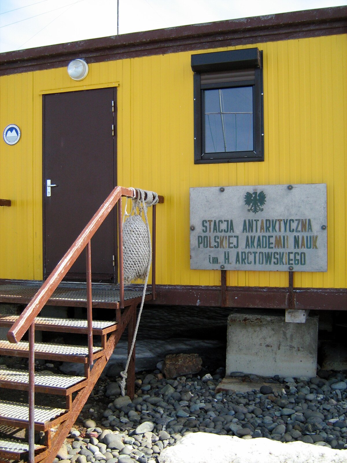 Zdjęcie przedstawia drzwi wejściowe do budynku stacji badawczej. Budynek znajduje się około metra nad ziemią, prowadzą do niego metalowe schody. Obok drzwi tablica z napisem „Stacja antarktyczna Polskiej Akademii Nauk imienia Henryka Arctowskiego”. Nad napisem godło Polski.