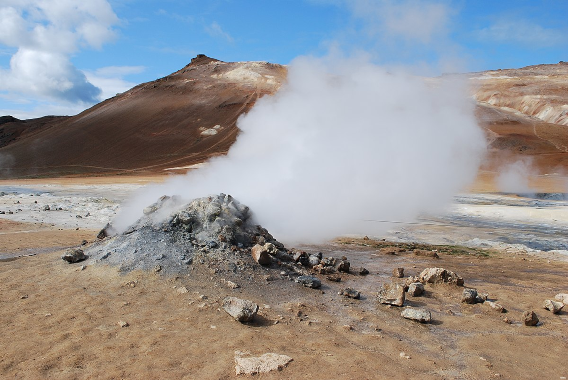 Grafika przedstawia zdjęcie fumaroli, czyli wyziewu wulkanicznego (szczeliny w skorupie ziemskiej, która jest objawem aktywności wulkanicznej), z którego wydobywa się biały gaz. Znajduję się on u podnóża brązowej góry. Na pierwszym planie widoczny jest brązowy piasek i szary krater w ziemi, z którego wydobywa się gaz.