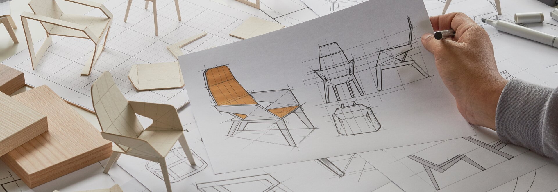 Fotografia przedstawia stół, na którym rozłożone zostały materiały oraz kartki z różnymi rzutami projektowanego krzesła o plastikowej ramie i pomarańczowym obiciu. Z lewej strony stołu leżą prostokątne kawałki drewna oraz wzornik wykonany z grubej tektury. Widoczne są projekty krzesła w kolorze jasnego drewna oraz dłoń mężczyzny trzymająca ołówek oraz kartkę z głównym projektem w centrum fotografii.