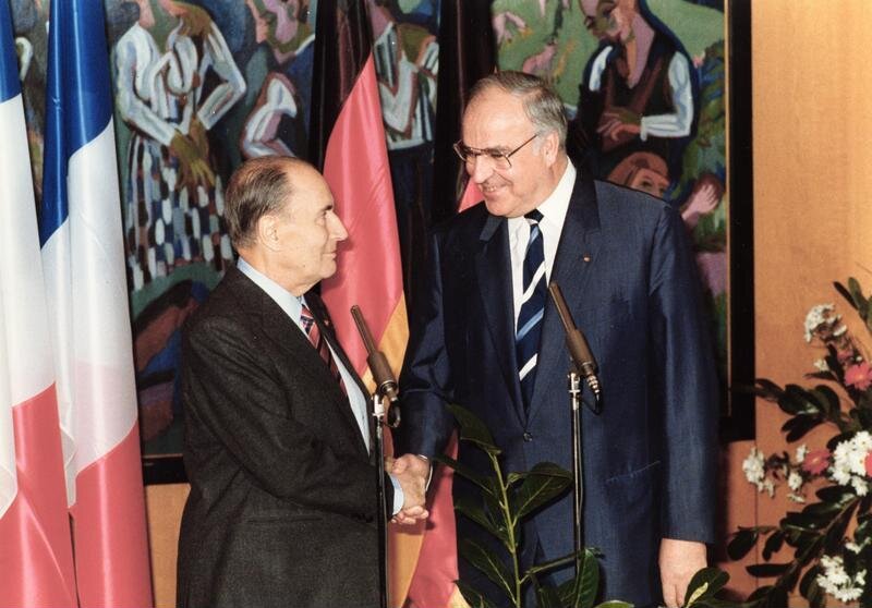 Zdjęcie przedstawia dwóch mężczyzn w garniturach ściskających sobie dłonie. Stoją przed mikrofonami. W tle widoczne są flagi Niemiec oraz Francji.  