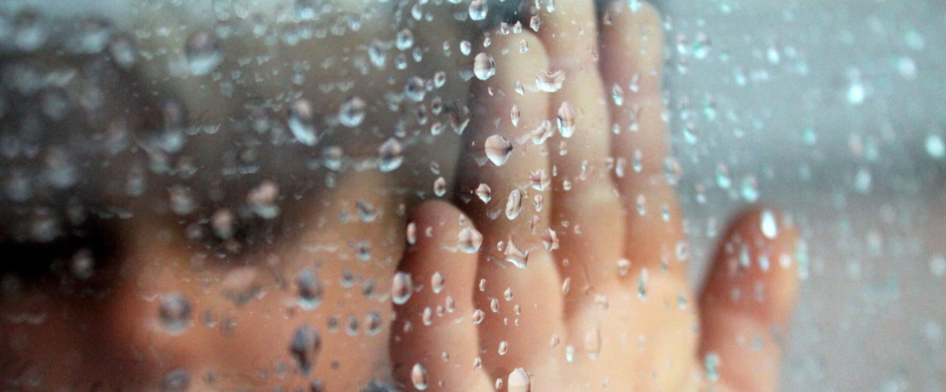 Zdjęcie przedstawia dłoń za szybą, na której znajdują się krople deszczu.