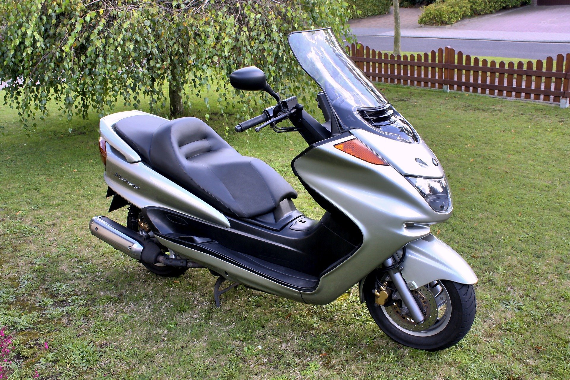 Zdjęcie przedstawia skuter. Jest srebrny. Ma dosyć długą przednią szybę, kanciasty tył. Stoi na trawie. 