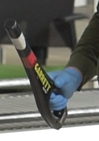 Zdjęcie przedstawia HHMD, czyli ręczne urządzenia do wykrywania metalu. Na zdjęciu widać rękę w niebieskiej rękawiczce lateksowej trzymającą urządzenie. Ma ono kształt prostego pręta, który płasko rozszerza się trochę u dołu. W miejscu rozszerzenia jest otwór, tworząc uchwyt urządzenia.