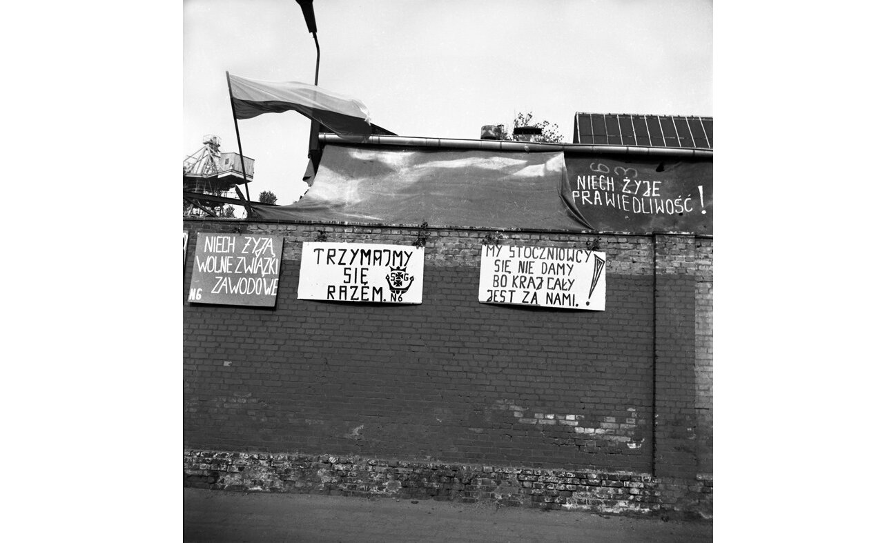 Czarno-biała fotografia przedstawiająca transparenty wywieszone na murze. Znajdują się na nich hasła "niech żyją wolne związki zawodowe", "trzymajmy się razem", "my stoczniowcy się nie damy, bo kraj cały jest za nami" czy "niech żyje sprawiedliwość".