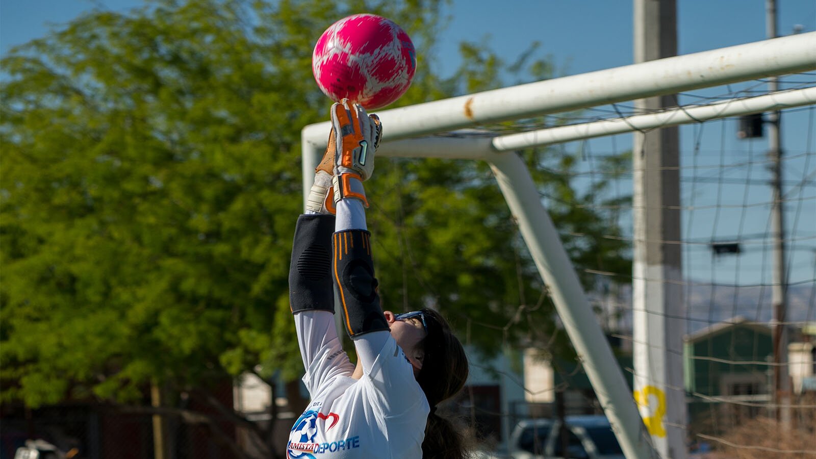Zdjęcie przedstawia dziewczynę grającą na pozycji bramkarza. Ujęcie przedstawia dziewczynę od pasa w górę, w wyskoku, łapiącą piłkę lecącą w okno bramki. Dziewczyna ubrana jest w bluzkę z długim rękawem, na dłoniach ma rękawice bramkarskie. Po prawej stronie widoczna jest bramka. 