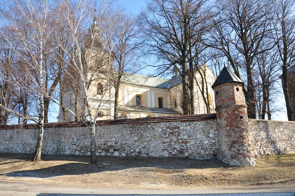 Zdjęcie przedstawia kościół otoczony kamiennym, jasnym murkiem. Wokół kościoła rosną drzewa. 