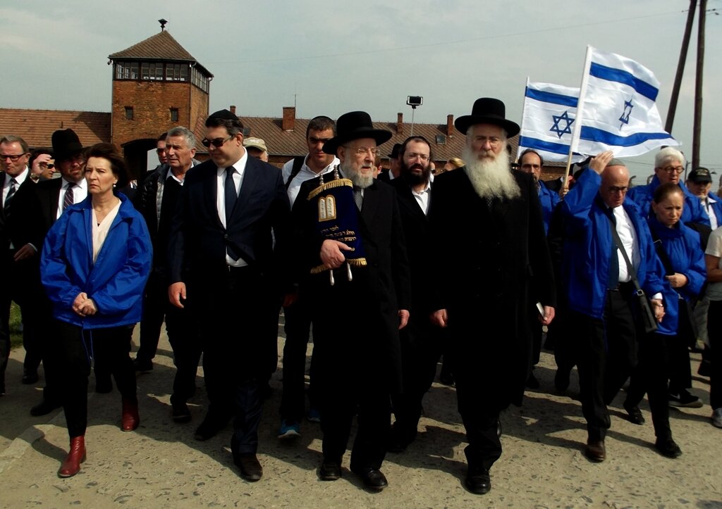 Fotografia barwna przedstawiająca uczestników Marszu Żywych. Są wśród nich mężczyźni w kapeluszach i jarmułkach, jeden z nich niesie Torę. Inni uczestnicy niosą flagi Izraela.