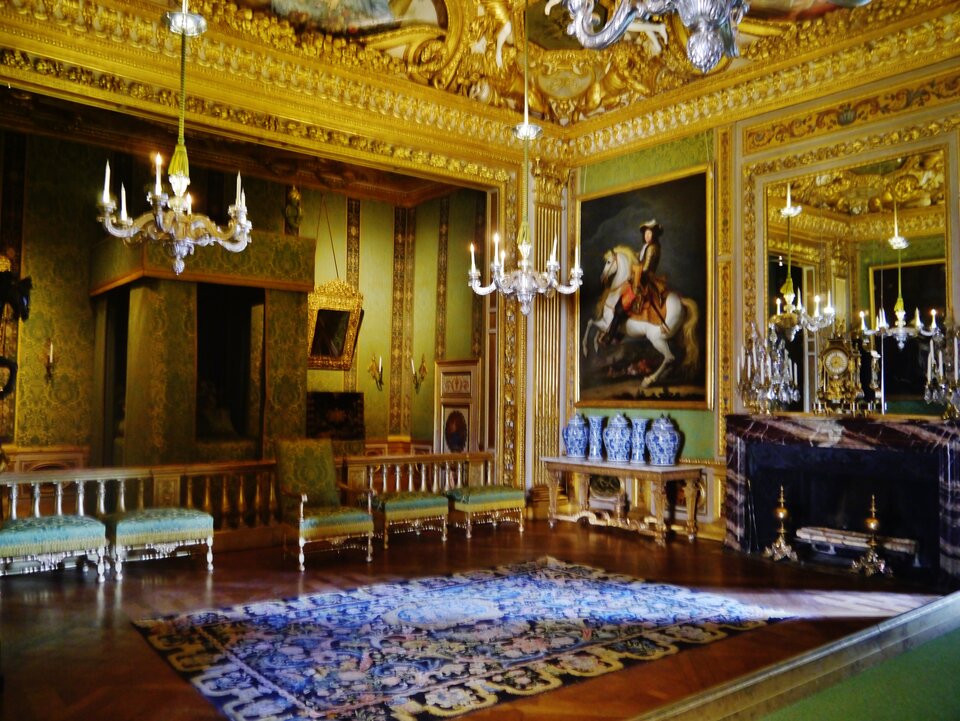 Bogato zdobione wnętrze pałacowej sali w stylu barokowym. Ściany pokryte są złoconymi ornamentami i zielonymi tkaninami, a z sufitu zwisają kryształowe żyrandole. W pomieszczeniu znajdują się eleganckie krzesła, kominek z marmurową obudową, duże lustra oraz obraz przedstawiający jeźdźca na koniu. Na podłodze leży ozdobny dywan.