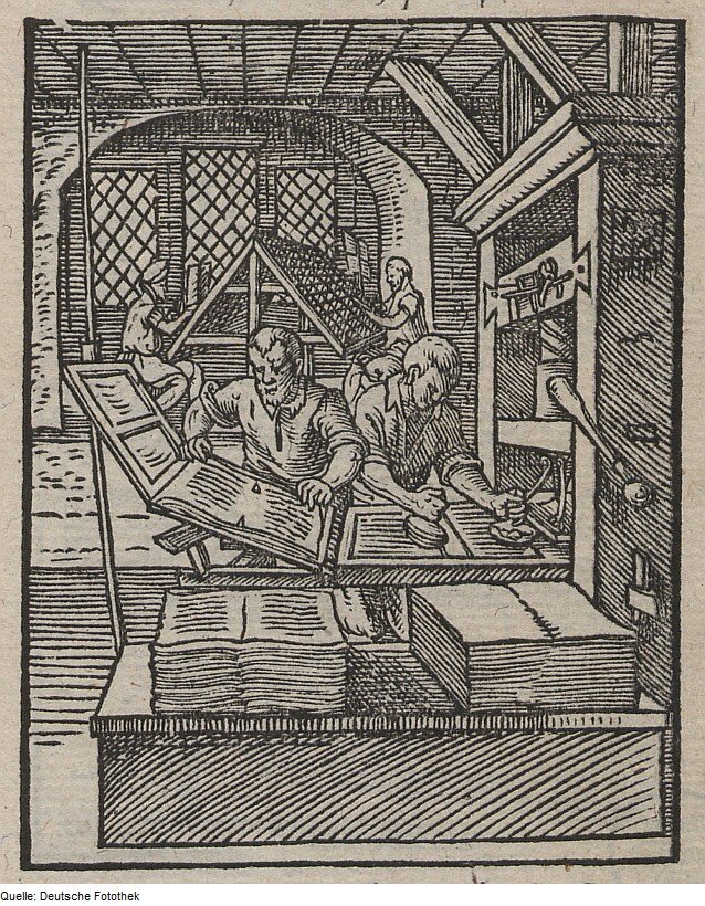 Rycina przedstawia czterech mężczyzn pracujących przy składaniu książek. Dwaj z nich nakładają farbę na czcionki. Są widoczni w oddali. Dwaj odbijają tekst na papierze. Przed nimi leżą w dwóch rzędach kartki. Te po lewej są zapisane, po prawej są czyste. Na ścianie po prawej stronie wiszą narzędzia drukarskie. 