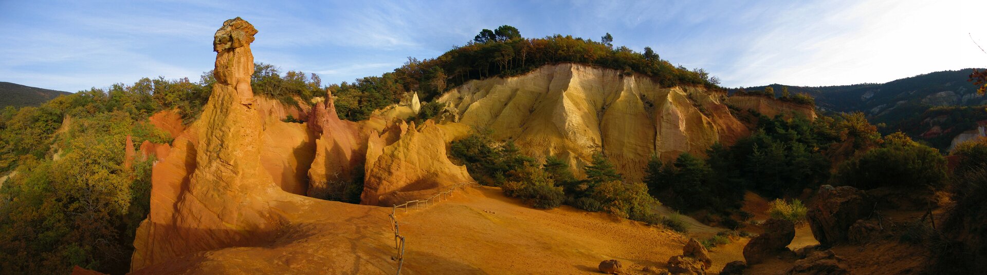 Na zdjęciu znajduje się panorama ukazująca skały w kolorze czerwono-brązowym. Są one pozbawione roślinności. Drzewa rosną po prawej i po lewej od skał. Nad skałami niebo jest jasne.