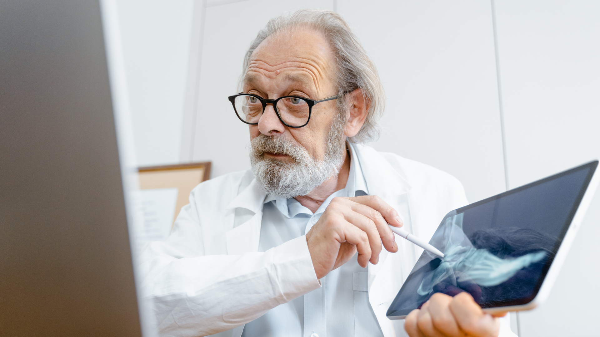 Zdjęcie przedstawia starszego, łysiejącego mężczyznę w okularach. Mężczyzna ma siwe włosy i brodę. Ubrany jest w lekarski fartuch. W ręku trzyma tablet, na którym wyświetla się zdjęcie rentgenowskie stopy. W drugim ręku mężczyzna trzyma długopis, którym wskazuje na zdjęcie rentgenowskie.