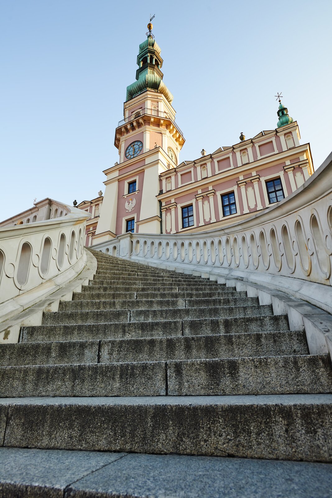 Ilustracja przedstawia zdjęcie ratusza w Zamościu od strony schodów. Ukazuje ujęcie z perspektywy żabiej, czyli od dolnej części schodów wejściowych do ratusza. Pierwszy plan zajmują dolne stopnie schodów z balustradą, które zwężają się w stronę ratusza. W górnej części widoczna jest elewacja frontowej części obiektu z masywną wieżą zegarową, zwieńczoną hełmem. Po prawej stronie elewacji znajdują się trzy okna, przedzielone dekoracją naścienną. Nad nimi front dekoruje wysunięta ścianka, zwieńczona rzeźbami - jest to attyka. Na brzegu znajduje się narożna wieżyczka, kryta hełmem.