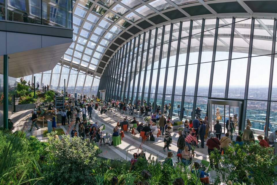 Ilustracja przedstawia Sky‑garden na jednym z poziomów Commerzbank Tower. We wnętrzu znajdują się liczne drzewka oraz krzewy. Budynek jest oszklony. W środku znajduje się mnóstwo ludzi.