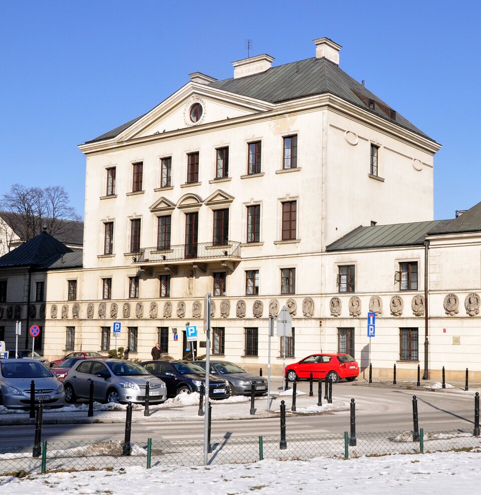 Na zdjęciu widać jasny, kilkukondygnacyjny budynek o prostokątnej bryle i symetrycznej elewacji. Fasada ma kremowy kolor, regularnie rozmieszczone prostokątne okna oraz centralny fragment z niewielkim balkonem z metalową balustradą. Dach jest ciemny, skośny, z kilkoma kominami. Na dolnej kondygnacji elewację zdobi pas okrągłych reliefów. Przed budynkiem znajduje się ulica oraz parking z zaparkowanymi samochodami. Przy ulicy ustawione są znaki drogowe, słupki oraz niskie ogrodzenie. Na chodniku i przy krawężnikach widoczny jest śnieg. W tle znajduje się bezchmurne, niebieskie niebo.