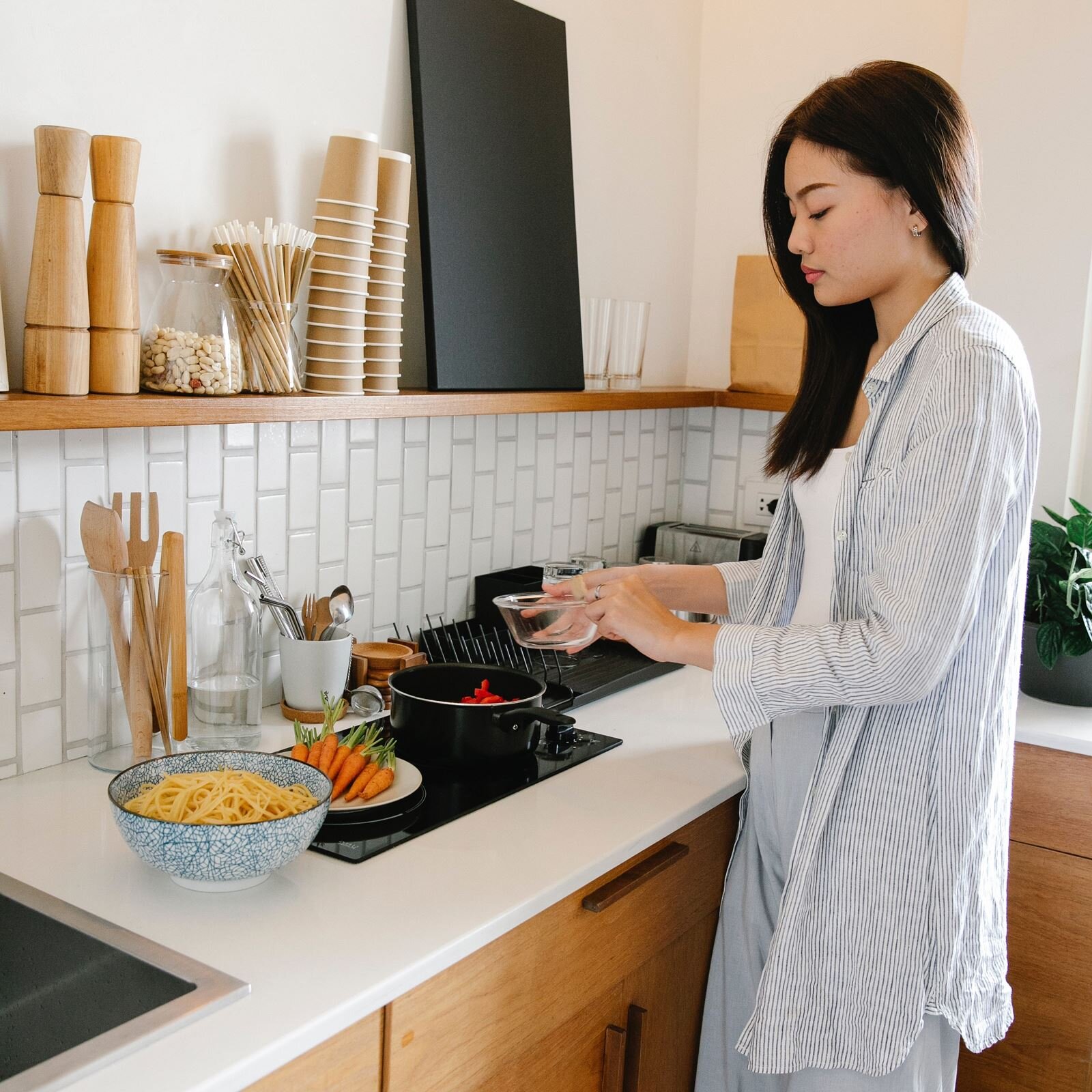 Zdjęcie przedstawia wnętrze kuchni, z długim blatem roboczym w kolorze białym, pod którym widoczne są drewniane szafki. Nad płytą indukcyjną, przy blacie, stoi młoda dziewczyna, która w ręku trzyma małą szklaną miseczkę nad czarnym garnkiem stojącym na płycie, w którym znajdują się pokrojone czerwone papryki. Obok na białym talerzu widoczne są oczyszczone marchewki. Na lewo, na blacie, widoczna jest głęboka misa z ugotowanym makaronem spaghetti. Na blacie kuchennym widoczne są drewniane akcesoria kuchenne, włożone do wysokiego szklanego pojemnika, butelka z wodą, biały kubek z różnymi narzędziami kuchennymi, drewniane podstawki, suszarka do naczyń oraz toster. Na drewnianej półce nad blatem widoczne są dwa drewniane młynki do przypraw, szklany pojemnik z ziarnami fasoli, szklany pojemnik z papierowymi słomkami, jednorazowe kubki, czarna tablica.