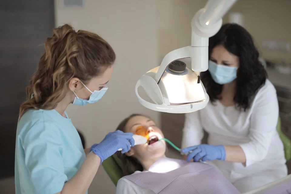 Zdjęcie przedstawia gabinet stomatologiczny. Na fotelu leży kobieta. Nad nią wisi lampa. Obok niej znajdują się kobieta stomatolog oraz pielęgniarka stomatologiczna. Obydwie mają założone fartuchy oraz maseczki. Trzymają rurkę oraz narzędzie przy zębach pacjentki.