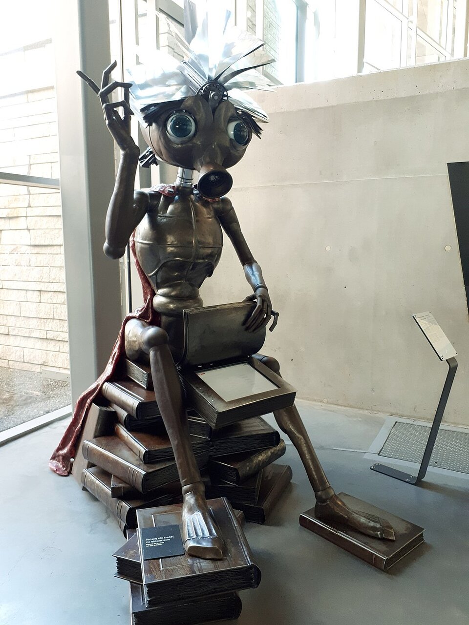 Fotografia przedstawiająca rzeźbę robota siedzącego na książkach. Robot otwiera książkę jedną dłonią, drugą unosząc do góry. 