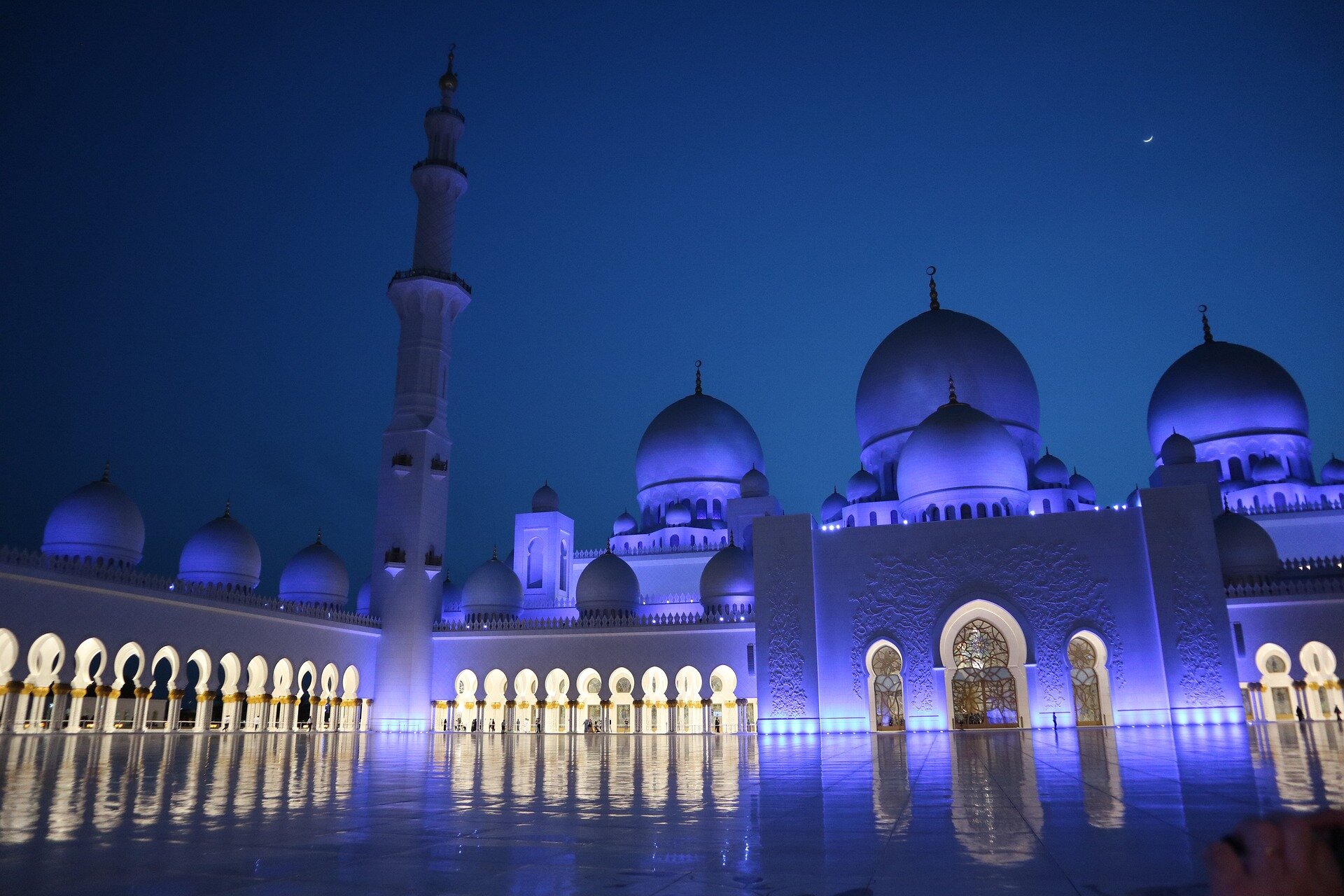 Zdjęcie przedstawia meczet w Abu Zabi w Zjednoczonych Emiratach Arabskich. Nocna fotografia przedstawia plac meczetu o błyszczącej powierzchni, w której odbija się budynek. Ma on białą fasadę, liczne kolumny połączone łukami, kopuły na dachu oraz strzelistą wieżę wznoszącą się wysoko ponad bryłę budynku. 