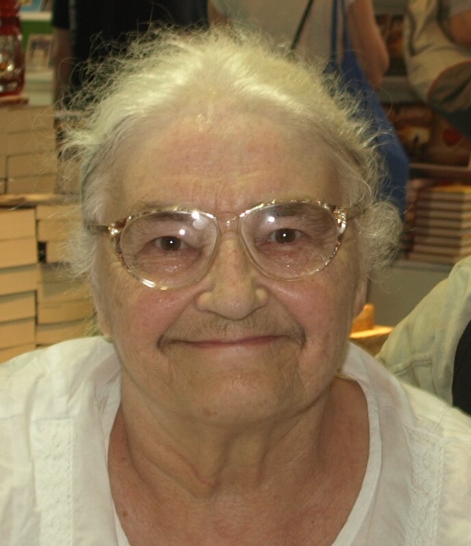 Fotografia przedstawia starszą kobietę. Ma białe włosy, okulary, krzaczaste, czarne brwi, pod nosem lekki zarost, delikatnie się uśmiecha. Jej wzrok jest wpatrzony prosto w widza. Ma na sobie białą bluzkę. W tle widać stosy książek i plecy osoby z niebieską torbą na ramieniu.