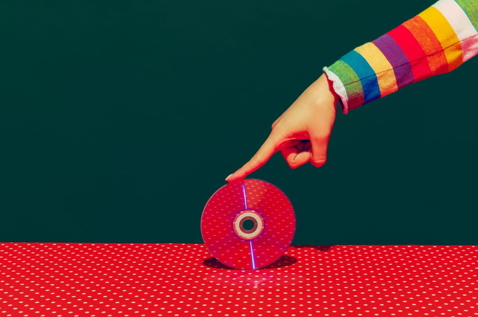 To fotografia aranżowana. Pokazuje rękę, która dotykając jednym palcem toczy płytkę CD po wzorzystym stole. Stół jest pokryty wzorem w kropki, który odbija się w lustrzanej powierzchni płytki. Dodatkowo wyciągnięta ręka ubrana jest w pasiasty sweter, w kolorach ciepłych, gorących i zimnych. Jest to z pewnością fotografia artystyczna.