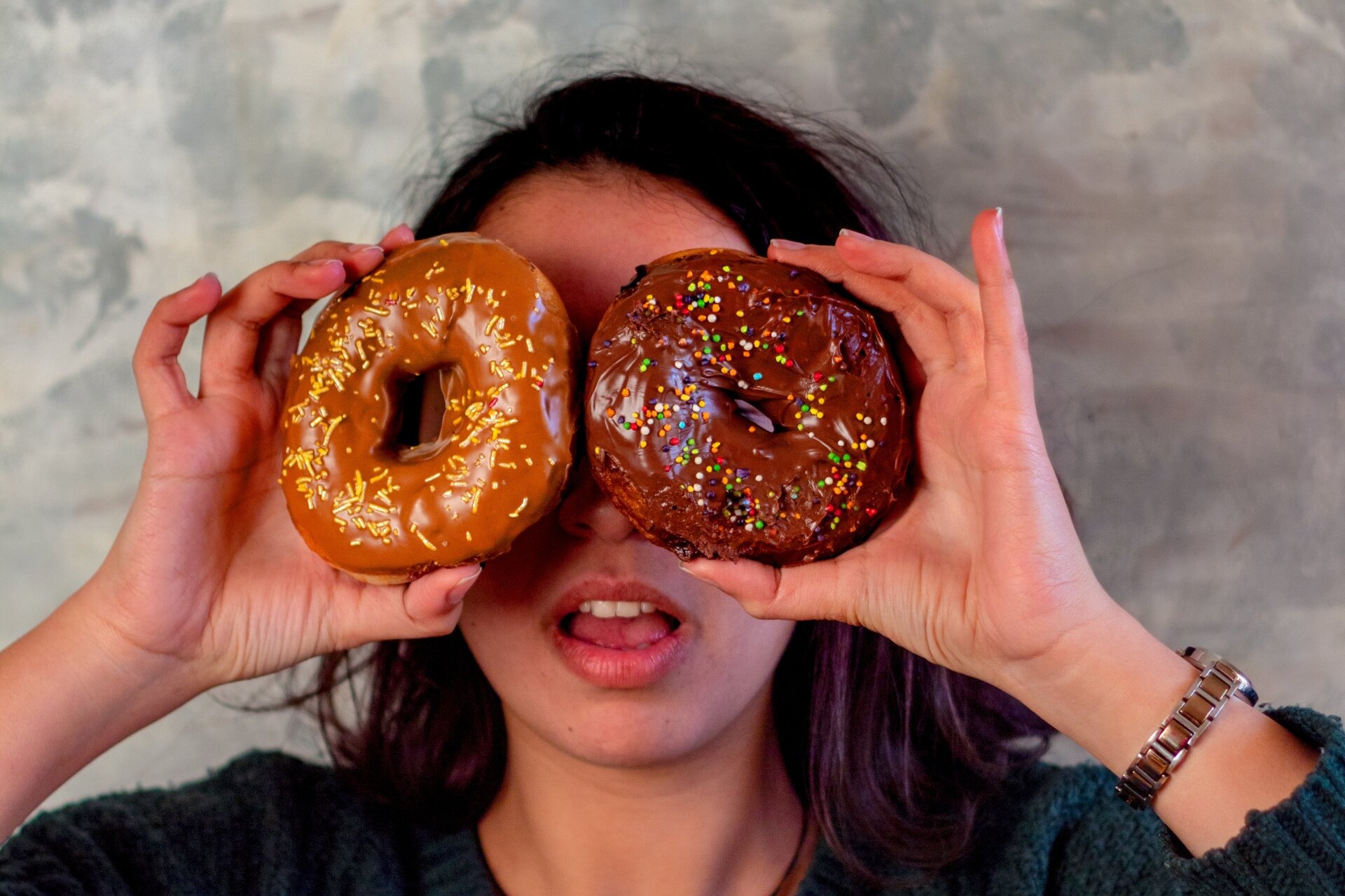 Zdjęcie przedstawia kobietę. Trzyma dwa donaty, zasłaniając sobie nimi oczy. Oba ciastka są z polewą i kolorową posypką.