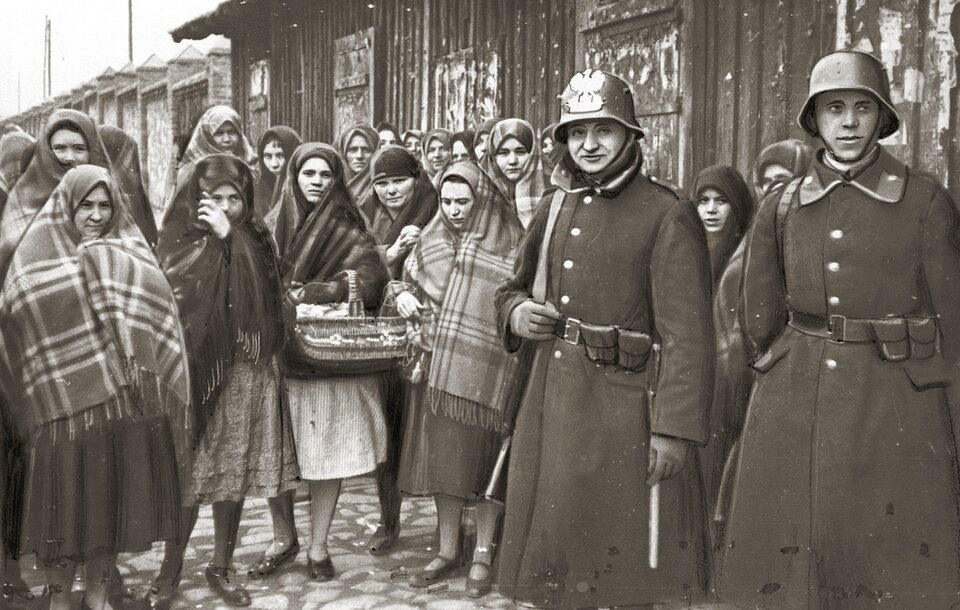 Zdjęcie przedstawia grupę kobiet. Stoją na brukowanej ulicy. Na pierwszym planie stoją dwaj żołnierze. Mają na sobie płaszcze mundurowe z postawionymi kołnierzami. Na głowie mają hełmy. Kobiety stojące z lewej strony i na środku są okryte chustami w kratę. Mają spódnice i płaskie buty. Jedna z nich trzyma pleciony koszyk. W tle jest budynek.