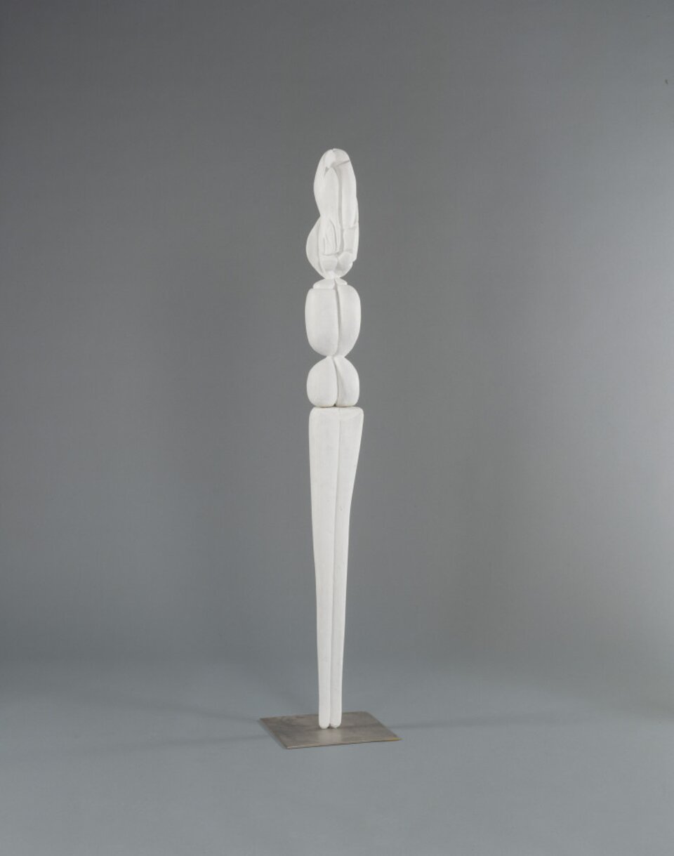 Zdjęcie przedstawia długą białą rzeźbę, na której znajdują się trzy białe kulki. Tło ilustracji jest szare.