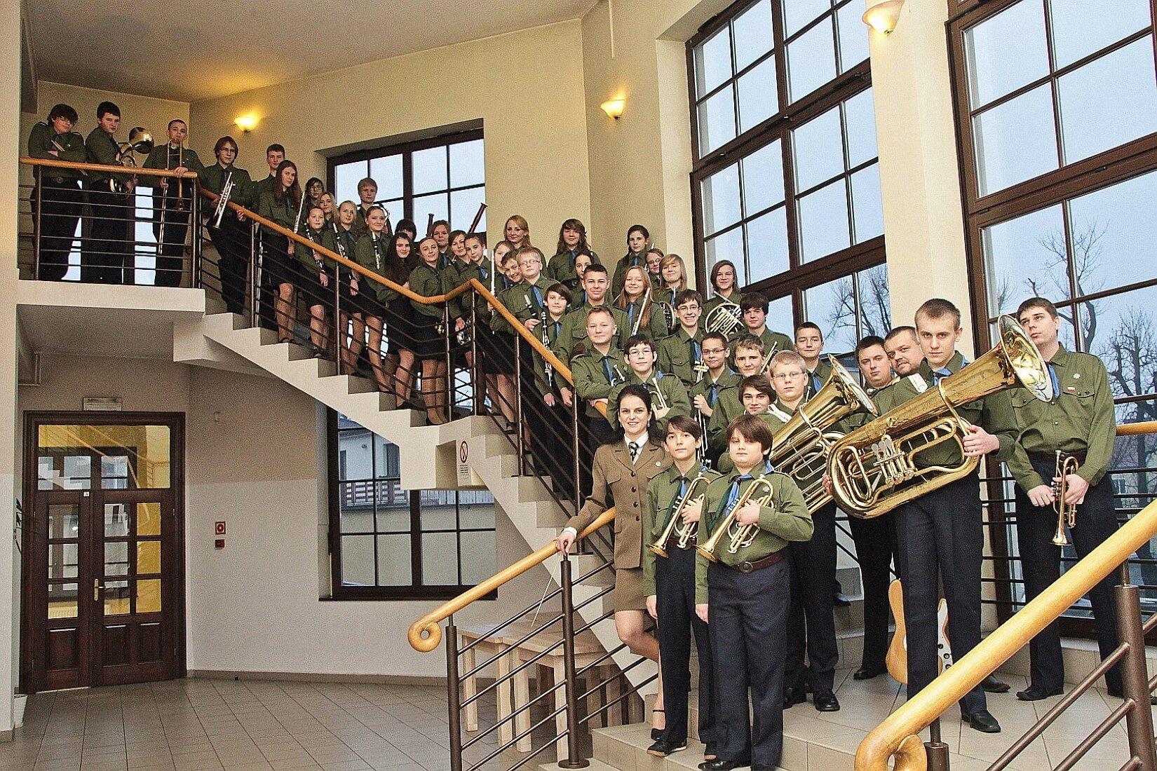 Zdjęcie przedstawia Harcerską Orkiestrę Dętą. Na zdjęciu znajdują się schody, na których stoją osoby ubrane w harcerskie mundury. Niektóre z osób trzymają w rękach różnego rodzaju instrumenty dęte. W tle zdjęcia znajduje się pomieszczenie z dużą ilością okien.