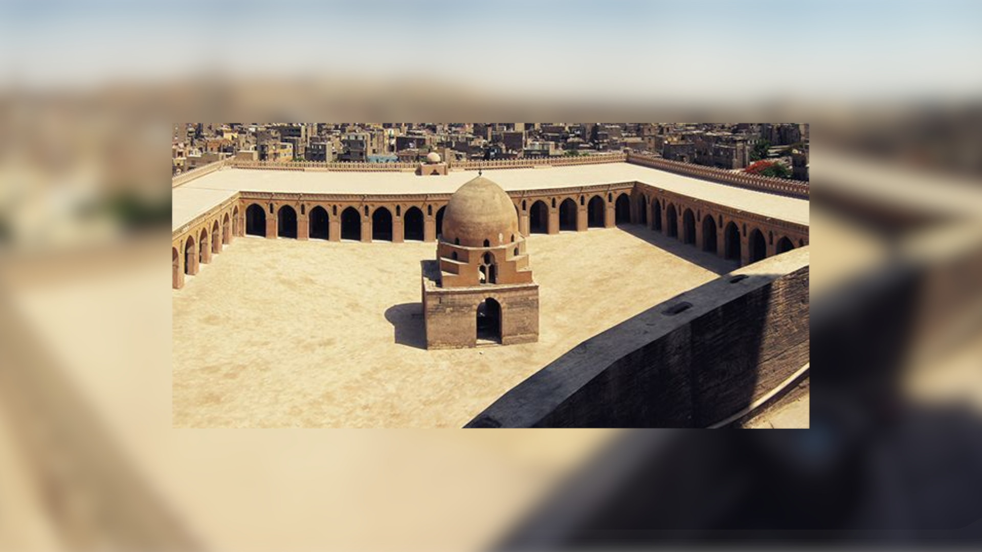 Na zdjęciu znajduje się dziedziniec meczetu, na którego środku znajduje się budowla z kopułą. Ściany meczetu składają się z łuków. W oddali znajdują się liczne budynki miasta. 